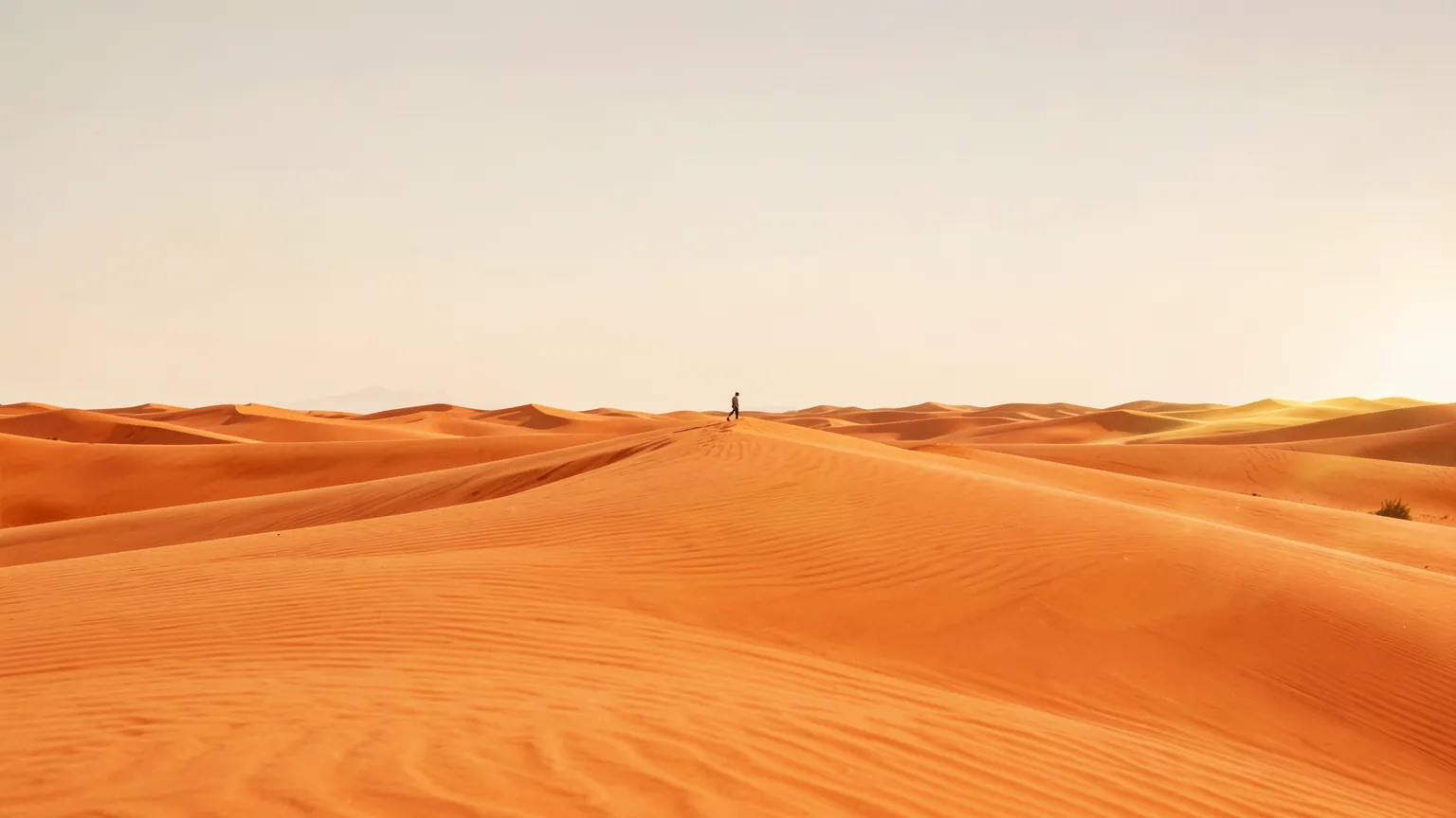Enorme paisaje desértico de dunas anaranjadas bajo un cielo brumoso, con una figura diminuta caminando por la cresta de una duna