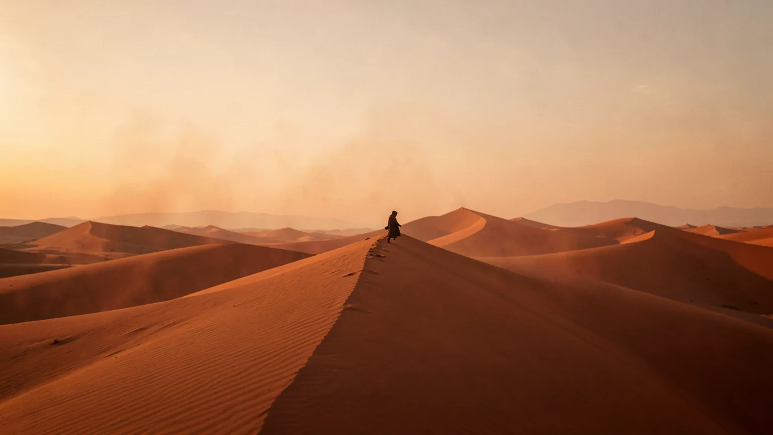 Desierto de dunas anaranjadas al atardecer con una figura solitaria caminando por la cresta bajo un cielo brumoso