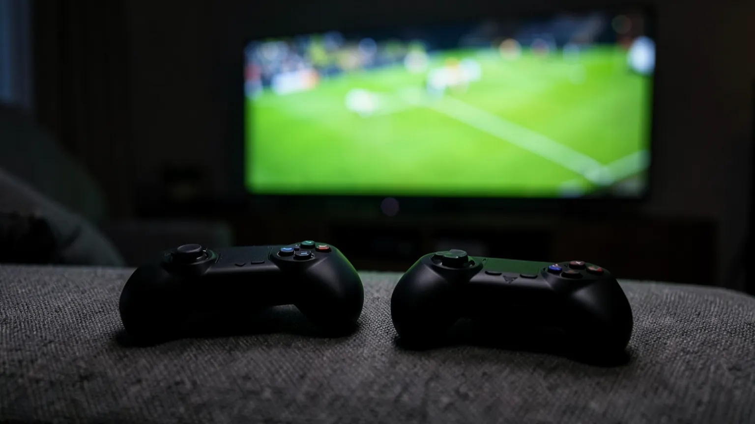 Dos mandos de consola sobre un sofá frente a un televisor encendido en un salón a oscuras